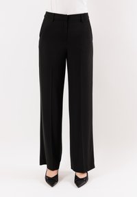 Pantaloni neri a gamba larga realizzati in tessuto liscio, con una parte frontale piatta, passanti per la cintura e tasche laterali. Abbinati a tacchi neri a punta.
