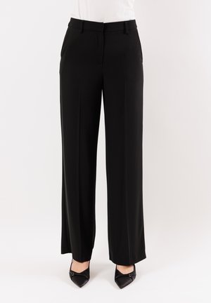 Pantaloni neri a gamba larga realizzati in tessuto liscio, con una parte frontale piatta, passanti per la cintura e tasche laterali. Abbinati a tacchi neri a punta.