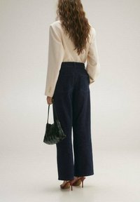 Mujer con cabello rizado y largo, vistiendo una blusa de color crema, jeans oscuros de pierna ancha y cintura alta, sandalias rojas de tacón, sosteniendo un bolso negro de tejido, de espaldas.