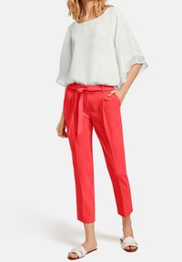 Chemisier blanc ample avec des manches évasées en voile, associé à un pantalon corail court à taille nouée. Les chaussures sont des sandales blanches.