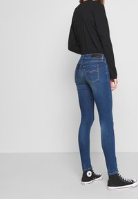 Jeans ajustados de mezclilla azul oscuro con cremallera, cinco bolsillos y costuras en contraste. Combinados con zapatillas negras de caña alta.