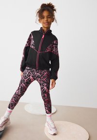 Black zip-up track jacket with pink graphic design, paired with black leggings featuring pink abstract patterns, and light sneakers with detailing.