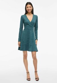 Teal, ribbed long-sleeve dress with V-neckline, fitted waist, and flared skirt. Worn with black strappy heels, emphasizing the dress's flow.