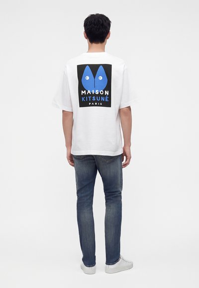 Άνδρας με γυρισμένη πλάτη φοράει λευκό T-shirt Maison Kitsuné με μπλε λογότυπο αλεπούς, συνδυασμένο με μπλε τζιν και λευκά αθλητικά παπούτσια, σε απλό φόντο.