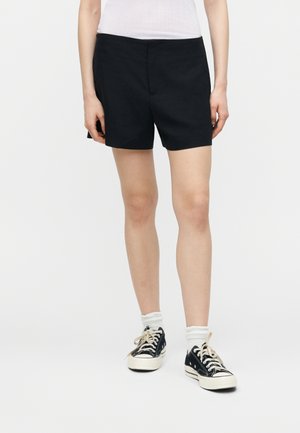 Person iført sorte shorts, hvide sokker og sorte lave sneakers, stående mod en ensfarvet hvid baggrund.