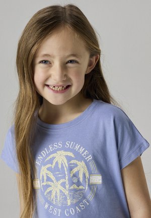 Niña sonriente con cabello largo castaño claro que lleva una camiseta lila con una palmera y el texto "Verano infinito Costa Oeste".