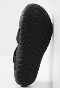 Zapato negro con una suela de goma texturizada, que presenta un patrón ondulado y correas ajustables. El material superior es liso, con un diseño minimalista.