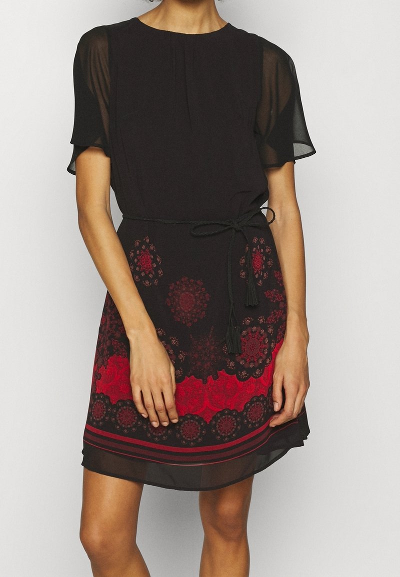 Femme portant une robe noire avec des manches courtes transparentes, des motifs floraux rouges et noirs sur la jupe, et une ceinture noire nouée à la taille.