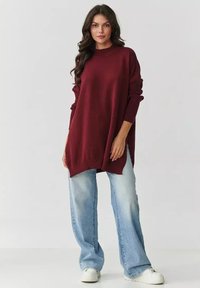 Bordowy oversize'owy sweter dzianinowy z prążkowanymi mankietami i dołem, w połączeniu z jasnoniebieskimi spodniami dzwonami i białymi trampkami, na neutralnym tle.