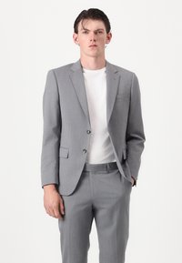Costume gris clair en tissu texturé avec une veste à un bouton et un pantalon à plis plats. Porté sur un t-shirt blanc.