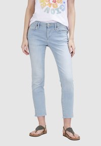 Jeans slim bleu clair avec un ourlet effiloché, cinq poches et des détails de bouton en métal. Portés avec un T-shirt graphique coloré et des sandales.