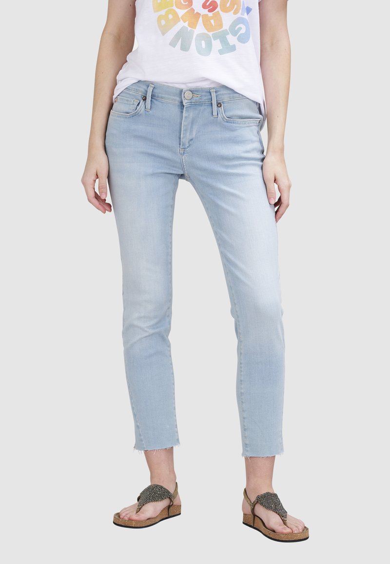 Jeans slim bleu clair avec un ourlet effiloché, cinq poches et des détails de bouton en métal. Portés avec un T-shirt graphique coloré et des sandales.