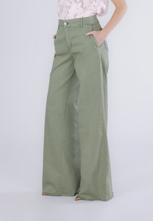 Donna che indossa pantaloni verde oliva a vita alta e gamba larga con orli sfrangiati, abbinati a una top in pizzo rosa chiaro, mani in tasca.