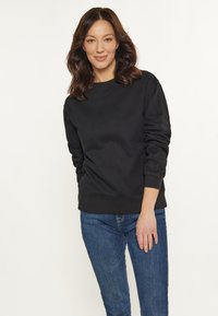 Fekete pulóver hosszú ujjal, kerek nyakkivágással és bordás mandzsettával, kék farmernadrággal párosítva. Puha tapintás és laza szabás.