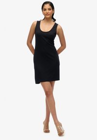 Robe noire sans manches en tissu doux, dotée d'un décolleté arrondi et d'une coupe décontractée. Portée avec des sandales transparentes à talons.