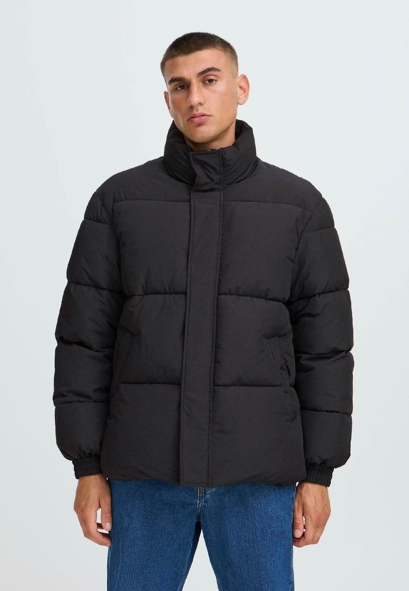 Νεαρός άνδρας που φοράει μαύρο μπουφάν puffer με ψηλό γιακά και μπλε τζιν, όρθιος μπροστά σε απλό ανοιχτό γκρι φόντο.