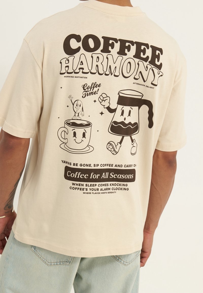 Beżowa bawełniana koszulka z czarnymi grafikami w stylu komiksowym przedstawiającymi filiżankę kawy i dzbanek. Tekst zawiera "Harmonia kawy" oraz wesołe frazy związane z kawą.