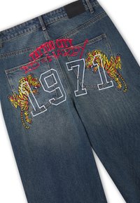 Dark blue denim jeans with embroidered text "TATTOO CITY ED HARDY 1971" and two yellow tigers on back pockets.
