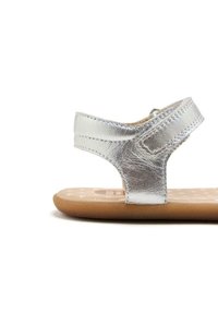 Sandalia metálica plateada con suela marrón plana, correa ajustable en el tobillo y textura suave. Presenta un diseño minimalista con costuras sutiles.