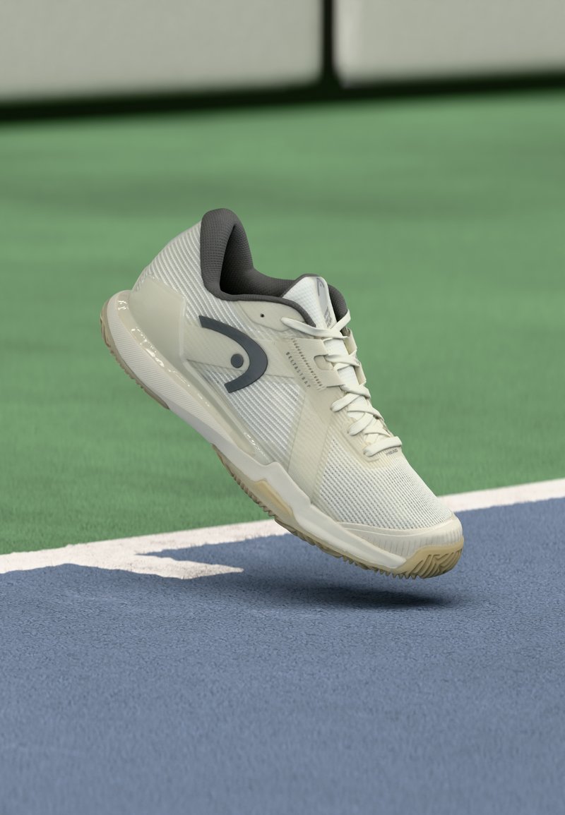 Ανοιχτόχρωμο παπούτσι τένις με υφασμάτινο άνω μέρος με υφή, γκρι λεπτομέρειες και λαστιχένια σόλα, τοποθετημένο πάνω σε επιφάνεια γηπέδου, δείχνοντας λεπτομέρειες του σχεδιασμού.