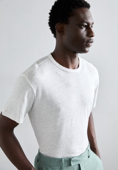 Kurzärmliges weißes T-Shirt aus leichtem Stoff mit Rundhalsausschnitt und subtiler Textur, kombiniert mit hellgrünen Hosen.