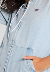 Light blue denim zip-up jacket with a hood, featuring a front pocket and a small logo patch. The fabric has a soft texture.