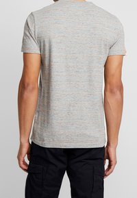 Muž oblečený v šedém tečkovaném tričku s krátkým rukávem a černých cargo kalhotách, zobrazený zezadu na jednolitém pozadí.