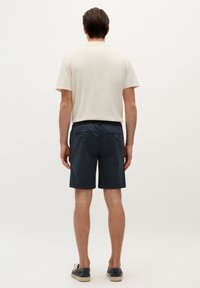 Beige kortærmet polo shirt og marineblå shorts med elastisk talje, der har to baglommer med knaplukninger, båret med mørke sko.