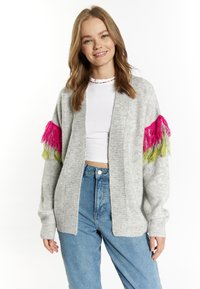 Cardigan grigio a maglia con frange rosa e verdi sulle maniche, design aperto frontale e polsini a coste. Indossato sopra una maglietta bianca e jeans.