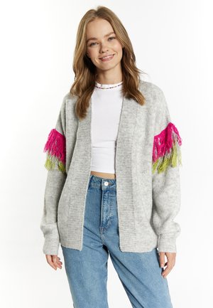 Harmaa neulecardigan, jossa on vaaleanpunaiset ja vihreät hapsut hihoissa, avonainen etuosa ja ribbireunat. Puettu valkoisen topin ja farkkujen päälle.