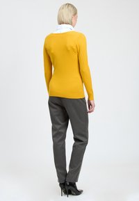 Helles gelbes Sweatshirt mit geripptem Saum, kombiniert mit maßgeschneiderten grauen Hosen und schwarzen High Heels, von hinten betrachtet.