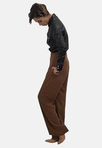 Pantaloni marroni a vita alta e gamba larga con una texture liscia, abbinati a una camicia in pelle nera fitted con bottoni a pressione e maniche lunghe.