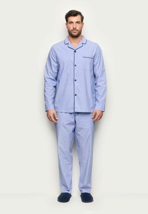 Mann steht in hellblauer, mit schwarzen Kanten versehener, geknöpfter Schlafanzugjacke und navyblauen Hausschuhen vor schlichten Hintergrund.