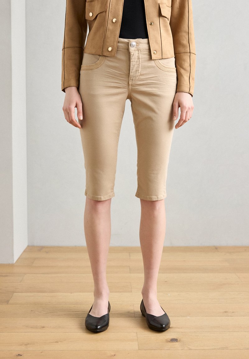 Beige bomullsshorts med en tettsittende design, som når rett over kneet. Parret med svarte leiligheter, plassert mot et lyst tregulv.