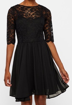 Schwarzes Kleid mit Spitzenoberteil und dreiviertel Ärmeln, ausgestattet mit einem fließenden, knielangen Rock aus transparentem Stoff.