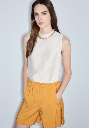 Jeune femme aux longs cheveux bruns portant un haut beige sans manches et un short jaune moutarde, les mains dans les poches, debout devant un fond clair uni.
