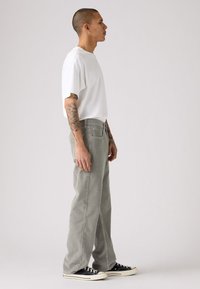 Man die zijwaarts staat in een wit T-shirt, grijze wijde broek, zwarte sneakers, en tatoeages op beide armen toont tegen een effen achtergrond.