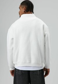 Witte oversized sweatshirt met een hoge kraag, geribbelde manchetten en een gestructureerde afwerking, gecombineerd met donkergrijze broek.