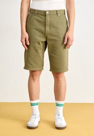 Personne portant un short vert olive jusqu'aux genoux, des baskets blanches et des chaussettes blanches avec deux bandes vertes, debout sur un sol jaune.