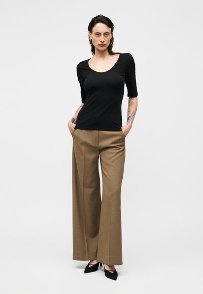 Γυναίκα με τατουάζ φοράει εφαρμοστή μαύρη μπλούζα με στρογγυλή λαιμόκοψη, καφέ παντελόνια με φαρδιά πόδια και μαύρες μυτερές γόβες, στέκεται με τα χέρια στις τσέπες.