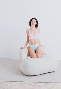Světle růžový bralette s bílým lemem a mátově zelenými kalhotkami, na krémovém, texturovaném, zakřiveném křesle s jemně zaoblenými okraji; neutrální pozadí.