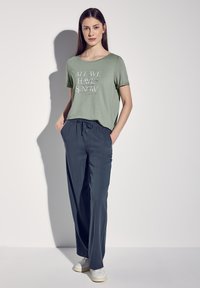 Jasnozielona koszulka z nadrukiem tekstowym, zestawiona z luźnymi, ciemnoszarymi spodniami na sznurek i białymi sneakersami.