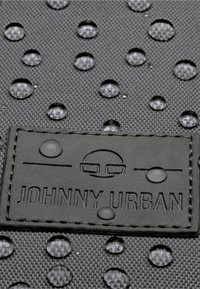 Fekete textúrájú anyag vízcseppekkel és egy fekete bőrcímkével, amelyen a "JOHNNY URBAN" felirat van domborítva. A cseppek mérete változó a felületen.