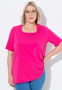 Top corto fucsia de manga corta con escote cuadrado y textura suave. Presenta un detalle de lado fruncido y un corte relajado. Se combina con pantalones azules.
