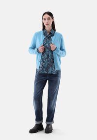 Hellblauer Cardigan über einem gemusterten dunkelblauen Hemd, kombiniert mit hochgekrempelten blauen Jeans und schwarzen Ankle Boots. Weicher Stoff und lockere Passform.