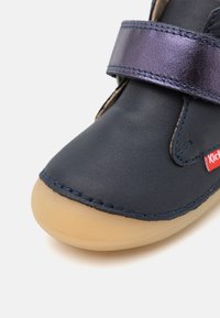 Chaussure en cuir marine avec une sangle velcro violette, bout arrondi et semelle en caoutchouc beige. Présente des coutures visibles et une étiquette de marque rouge.