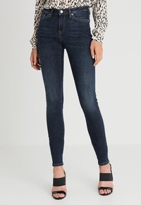 Mujer vistiendo jeans ajustados de color azul oscuro y sandalias de tacón alto negras y destalonadas, de pie contra un fondo claro y liso.