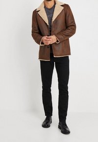 Brązowa skórzana kurtka z kremowym kołnierzem z owczej wełny, z zapięciem na guziki i bocznymi kieszeniami, noszona na szarym t-shircie i czarnych spodniach.