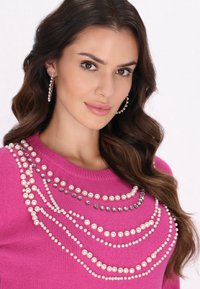 Maglione rosa lavorato a maglia con decorazioni a strati di perle e cristalli lungo il colletto. Il modello ha i capelli ondulati e indossa orecchini a cerchio.