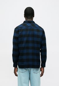 Camicia a quadri in flanella nera e blu, maniche lunghe, polsini con bottoni e colletto classico. Vista posteriore mostra l'orlo leggermente curvato.